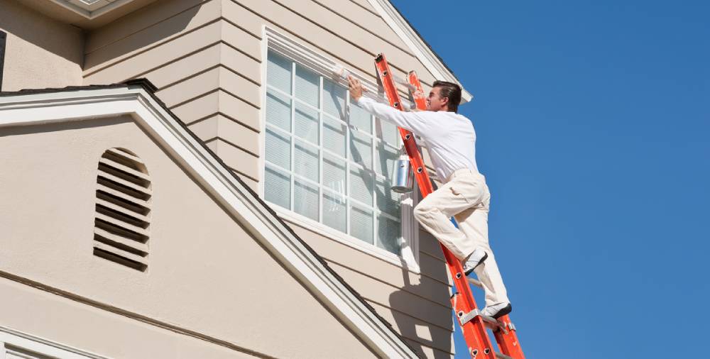 A side view of a male professional painter painting the exterior of a house against the heat of the sun.